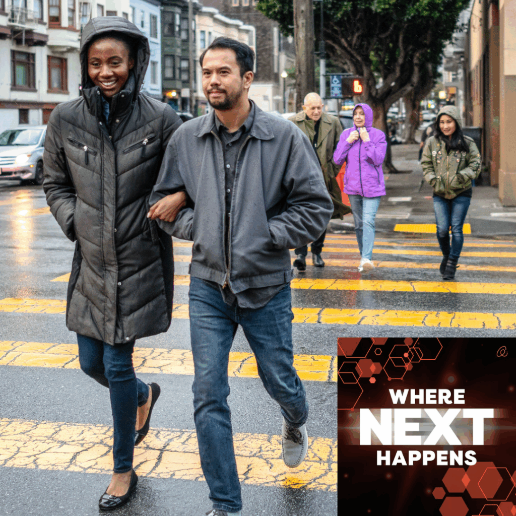 Two people in jackets walk arm in arm across a city crosswalk on a rainy day, with other pedestrians behind them and a text overlay reading “WHERE NEXT HAPPENS.”