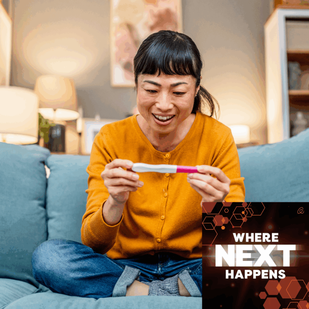 Woman sitting on a couch smiles while holding a home pregnancy test, with text overlay reading “WHERE NEXT HAPPENS.”